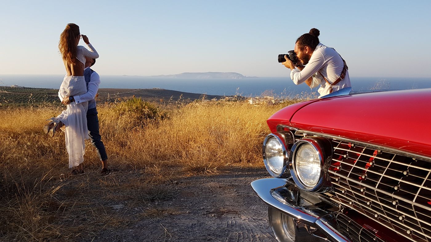 Φωτογράφος εν ώρα λήψης με την Cadillac σε πρώτο πλάνο και θέα στη θάλασσα